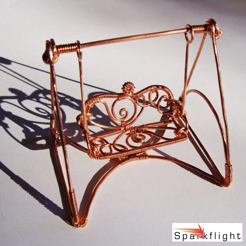 May include: A miniature copper wire swing with an ornate, heart-shaped backrest. The swing features intricate wire detailing and a decorative floral element at the top. The design includes curved supports and a horizontal bar for hanging.