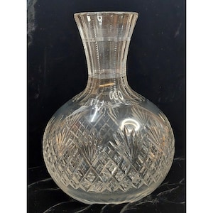 May include: A clear glass decanter featuring a rounded body and a flared neck. The surface is decorated with intricate cut patterns, including a diamond grid design and fan-shaped motifs that wrap around the lower portion of the vessel.