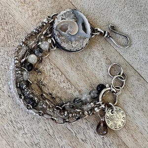 May include: A multi-strand bracelet featuring a large, round, gray and white stone centerpiece with a spiral design. The bracelet includes various chains, beads, and a small coin charm. The metal is silver-toned.