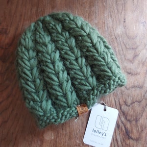 May include: A green knitted beanie with a cable knit pattern. The beanie has a small brown leather tag with the text "lolley's handmade" and the website "lolleys.co.uk".