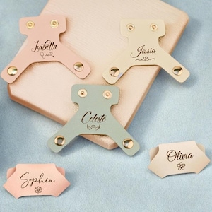 Könnte beinhalten: Eine Sammlung personalisierter Kabelorganisatoren aus Leder in verschiedenen Pastellfarben, darunter Rosa, Grün und Beige. Jeder Organizer hat einen Namen und dekorative Elemente und ist mit goldfarbenen Nieten befestigt.
