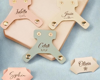 May include: A collection of personalized leather cable organizers in various pastel colors, including pink, green, and beige. Each organizer features a name and decorative elements, secured with gold-colored rivets.