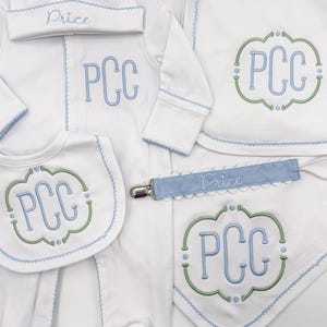 Könnte beinhalten: Ein weißes Baby-Kleidungsset bestehend aus einem Strampler, einer Mütze, einem Lätzchen und einem Spucktuch mit hellblauer und grüner Stickerei. Die Teile zeigen das Monogramm PCC und den Namen Price in blauer Schrift sowie einen Schnullerhalter.