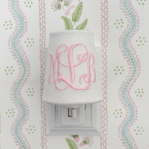 Puede incluir: Luz de noche blanca con una pantalla de tela con un monograma rosa. La luz de noche está enchufada a una toma de corriente blanca. El fondo es un papel pintado floral con detalles en rosa, verde y azul.