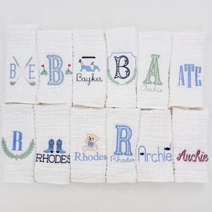 Puede incluir: Paños de muselina blancos para bebés con diseños bordados. Los diseños incluyen letras, palos de golf, un carrito de golf, un osito de peluche y botas de vaquero. El bordado es en tonos azules y verdes.