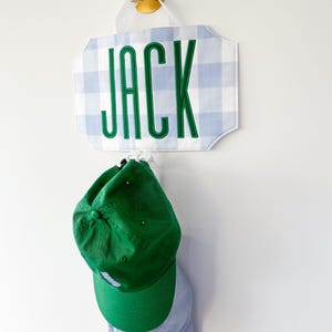 May include: A wall-mounted sign displaying the name "JACK" in green lettering, set against a blue and white gingham pattern. Beneath the sign hang a green and a light blue baseball cap.
