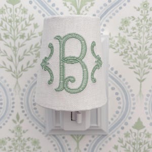 May include: A white night light with a fabric shade embroidered with a green letter "B". The night light is plugged into a white wall socket. The background is patterned wallpaper with green and blue floral designs.