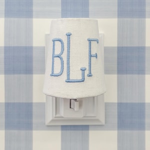 以下が含まれることがあります： ライトブルーの文字「BLF」が刺繍された布製シェードが付いた白いナイトライト。 ナイトライトは、青と白のチェック柄の壁に対して白いコンセントに差し込まれています。