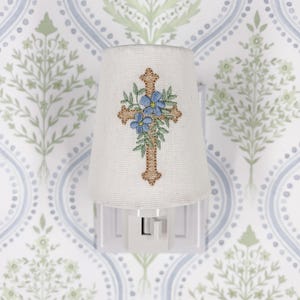 Op de afbeelding: Een wit nachtlampje met een stoffen kap met een kruisontwerp. Het kruis is bruin geborduurd met blauwe bloemen. Het nachtlampje is aangesloten op een muur met behang.