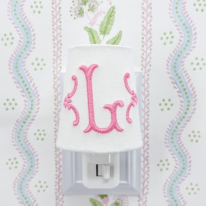 May include: White night light with a fabric shade. The shade features a pink embroidered letter "L" in a decorative font. The night light is plugged into a white wall outlet, set against a floral wallpaper background.