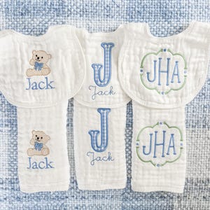 Puede incluir: Baberos y paños de eructo blancos para bebés con bordados azules y verdes. Un babero presenta un osito de peluche, mientras que otros muestran la letra "J" o las iniciales "JHA" y el nombre "Jack".