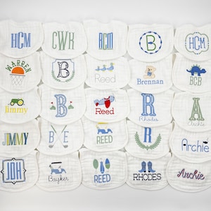 May include: A collection of white baby bibs with various embroidered designs. The bibs feature initials, names, and images like a basketball, tractor, airplane, and golf-themed elements. The embroidery colors include blue, green, and red.