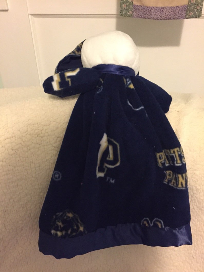 May include: A blue fleece baby blanket with a white head and a satin trim. The blanket has the Pittsburgh Panthers logo and the letters "PITT" and "PANTHERS" printed on it.