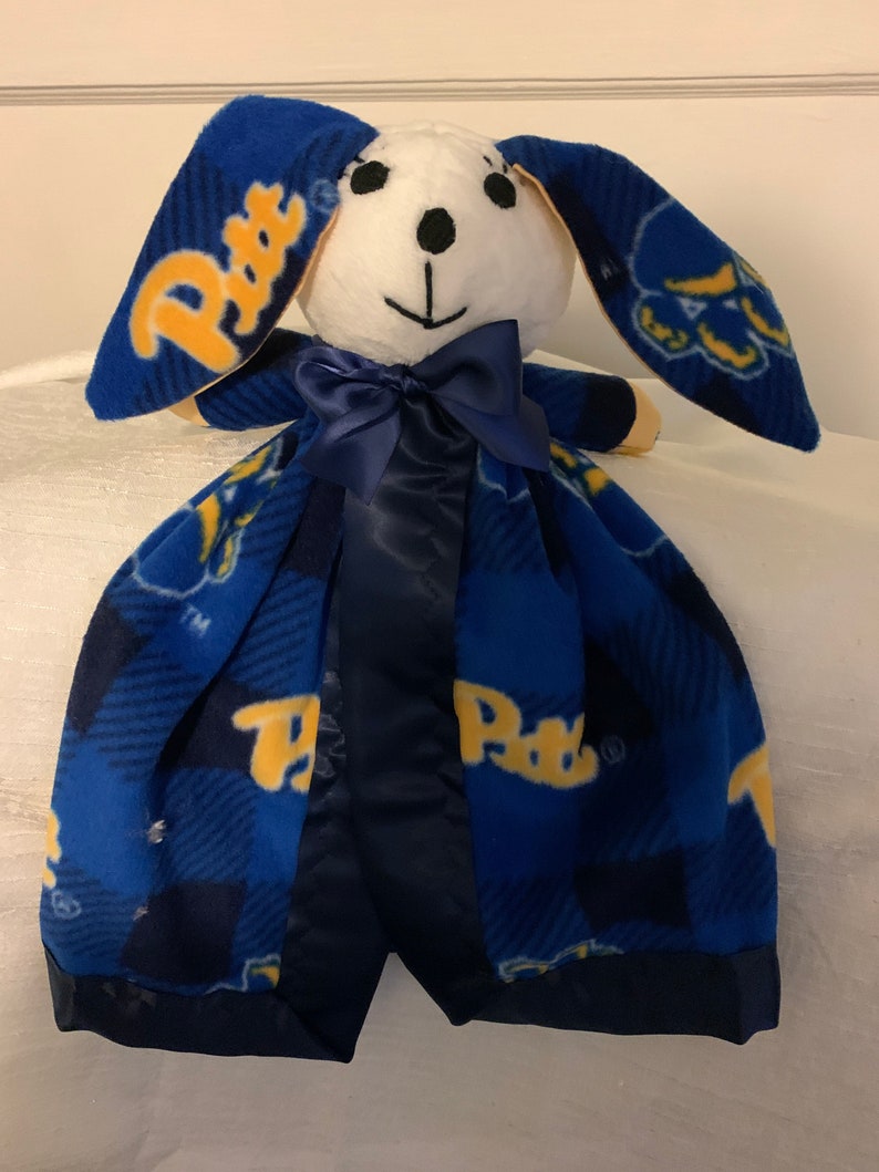 May include: A blue and yellow fleece baby blanket with a plush white bunny head attached. The bunny has black eyes and a black nose. The blanket has a blue and yellow plaid pattern with the word "Paws" repeated throughout.