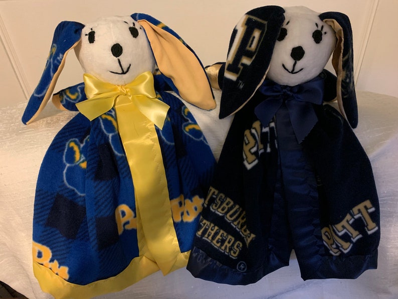 May include: Two plush bunny security blankets with a blue and gold plaid pattern. One blanket has a yellow ribbon and the other has a blue ribbon. Both blankets have the Pitt logo and the words "Pittsburgh Panthers" on them.