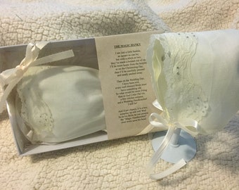May include: A white handkerchief and bonnet set, presented in a box with a poem. The bonnet features lace trim and small embellishments. A white ribbon is tied around the bonnet. The text on the card reads "THE MAGIC HANKY".
