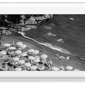 May include: A black and white aerial photograph of a beach with striped umbrellas and people relaxing on lounge chairs. The beach is bordered by a rocky cliff and the ocean.