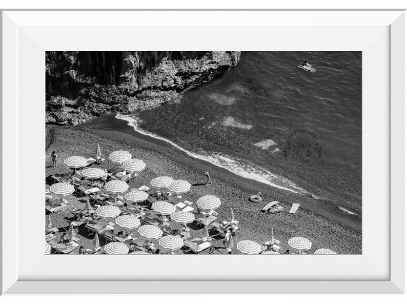 May include: A black and white aerial photograph of a beach with striped umbrellas and people relaxing on lounge chairs. The beach is bordered by a rocky cliff and the ocean.
