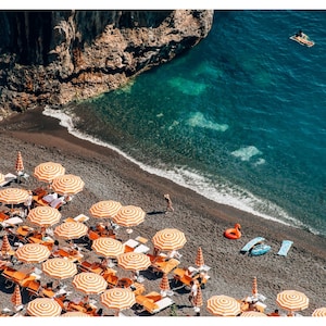 May include: An aerial view of a black sand beach with orange and white striped beach umbrellas. The beach is lined with lounge chairs and there are people swimming in the clear blue water.