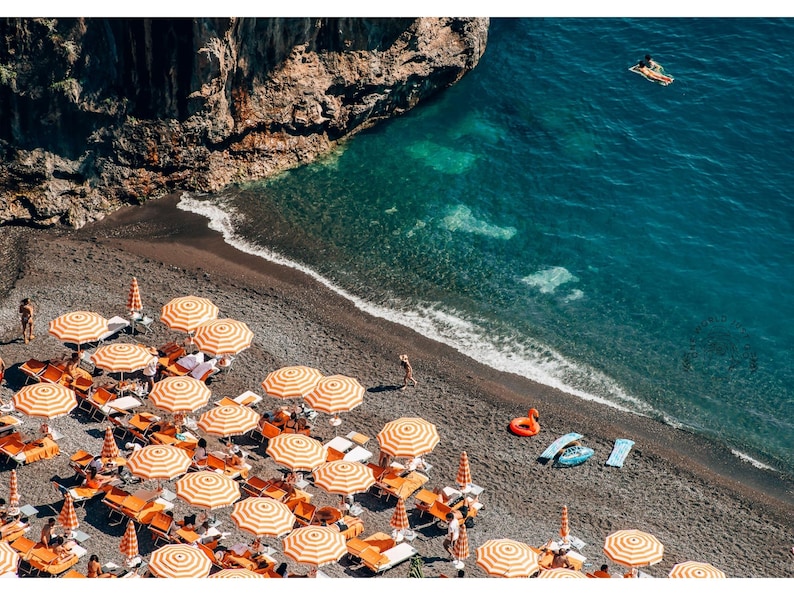 May include: An aerial view of a black sand beach with orange and white striped beach umbrellas. The beach is lined with lounge chairs and there are people swimming in the clear blue water.