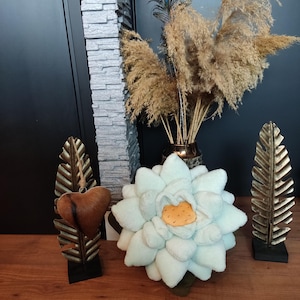 Op de afbeelding: Een lichtblauw pluche bloemenkussen met een geel centrum, twee gouden metalen decoratieve standaards met bruine hartvormige ornamenten en een vaas met gedroogd pampasgras.