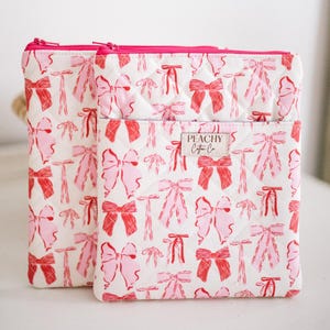 Könnte beinhalten: Zwei gesteppte Kosmetiktaschen mit weißem Hintergrund und einem sich wiederholenden Muster aus rosa und roten Schleifen. Die Taschen haben rosa Reißverschlüsse und den Text "PEACHY Cotton Co." auf einem Etikett.