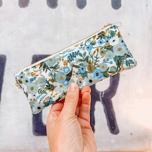 May include: A rectangular pencil case with a floral pattern in shades of blue, white, and gold. The case has a gold zipper and a small fabric tag. The case is held up by a hand.