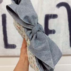 May include: A floral patterned clutch with a large gray bow. The clutch features a quilted design with a white background and small red, green, and pink flowers. The bow is made of a solid gray fabric and is tied in a knot.