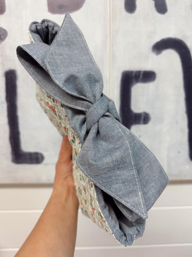 May include: A floral patterned clutch with a large gray bow. The clutch features a quilted design with a white background and small red, green, and pink flowers. The bow is made of a solid gray fabric and is tied in a knot.