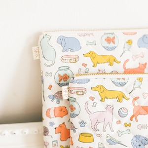 Könnte beinhalten: Zwei weiße, gesteppte Beutel mit einem farbenfrohen Muster zum Thema Haustiere. Das Muster enthält Illustrationen von Hunden, Katzen, Fischen, Vögeln und anderen Haustieren. Die Beutel haben goldene Reißverschlüsse.