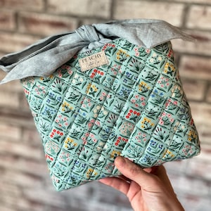 May include: A quilted, square-shaped bag with a floral pattern in shades of blue, yellow, and red on a mint green background. The bag is tied with a light blue fabric bow. The label reads "PEACHY Cotton Co."