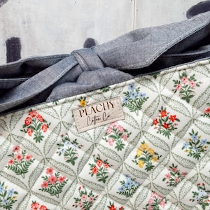 May include: Close-up of a quilted fabric with a floral pattern in red, pink, yellow, and blue. The fabric is white with a diamond pattern and green leaves. A denim-like fabric forms a bow. The label reads "PEACHY Cotton Co."
