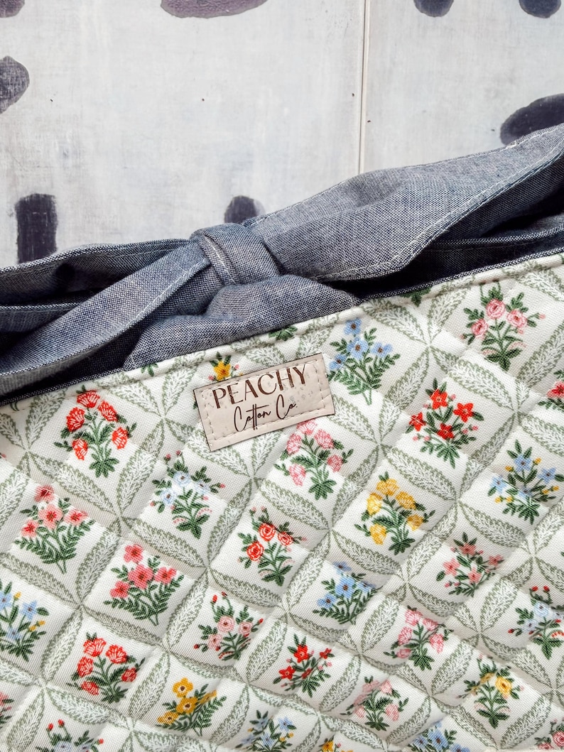 May include: Close-up of a quilted fabric with a floral pattern in red, pink, yellow, and blue. The fabric is white with a diamond pattern and green leaves. A denim-like fabric forms a bow. The label reads "PEACHY Cotton Co."