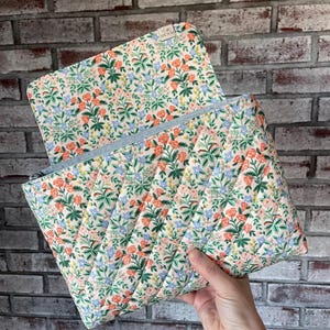 May include: Quilted floral print pouch with a light blue zipper. The pouch features a cream background with a pattern of orange, green, and blue flowers. The pouch is open, revealing the interior.