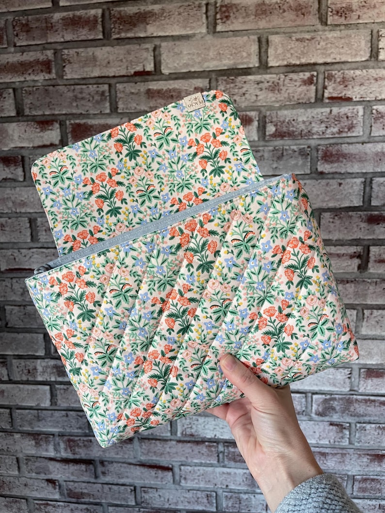 May include: Quilted floral print pouch with a light blue zipper. The pouch features a cream background with a pattern of orange, green, and blue flowers. The pouch is open, revealing the interior.