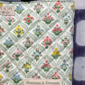 May include: Quilted fabric bag with a floral pattern in red, pink, yellow, and blue. The bag has a white background with a green leaf design. The text "PEACHY Cotton Co." and "Shannon S. Kennedy" are visible on the bag.