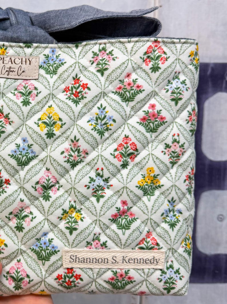 May include: Quilted fabric bag with a floral pattern in red, pink, yellow, and blue. The bag has a white background with a green leaf design. The text "PEACHY Cotton Co." and "Shannon S. Kennedy" are visible on the bag.