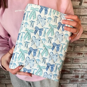 May include: Quilted white clutch with a repeating pattern of blue and teal bows. The clutch has a diamond pattern and a zipper closure. The words "MEETING YOU" are printed on the upper left side of the image.