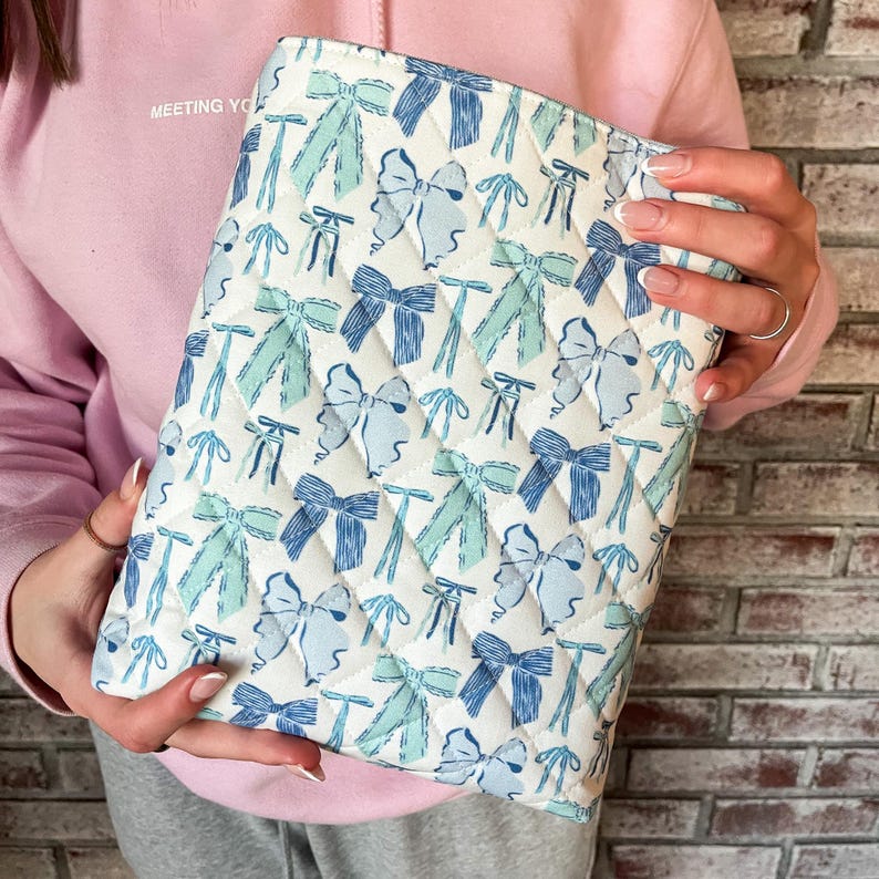 May include: Quilted white clutch with a repeating pattern of blue and teal bows. The clutch has a diamond pattern and a zipper closure. The words "MEETING YOU" are printed on the upper left side of the image.