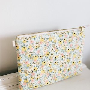 May include: A rectangular floral print pouch with a gold zipper. The pouch features a pattern of yellow, pink, and gold flowers on a white background. The zipper is gold-colored, and the pouch is sitting on a white surface.