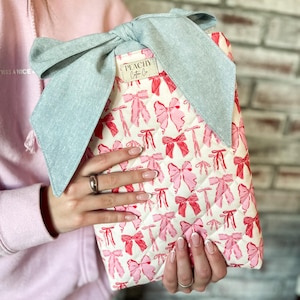 May include: A quilted fabric case with a repeating pattern of pink bows on a white background. The case is topped with a large, light blue bow. The label reads "PEACHY Cotton Co."