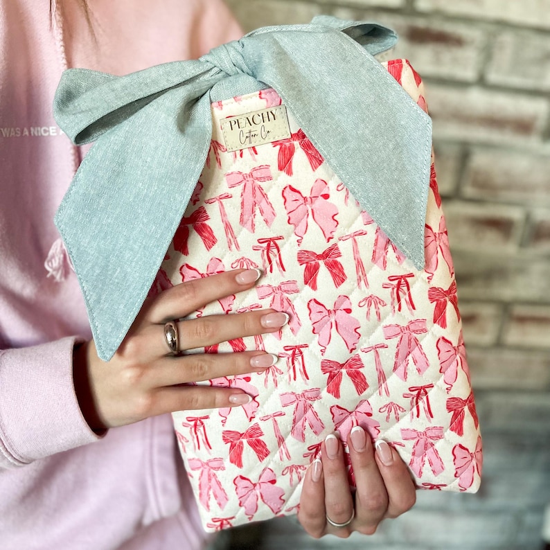 May include: A quilted fabric case with a repeating pattern of pink bows on a white background. The case is topped with a large, light blue bow. The label reads "PEACHY Cotton Co."