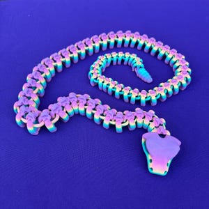 May include: A colorful, articulated snake toy in shades of purple, yellow, and blue. The snake is made of connected segments and has a stylized head and tail. The toy is displayed on a solid blue background.
