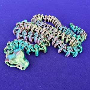 May include: A 3D-printed articulated snake toy in iridescent colors of green, purple, and gold. The snake is posed on a solid blue background. The head is detailed with eye and nostril features. The body is segmented, allowing for flexible movement.