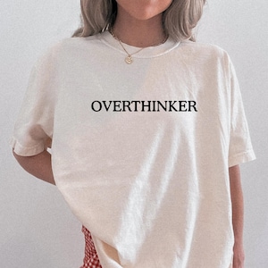 May include: A cream-coloured short-sleeve t-shirt featuring the word OVERTHINKER printed in black capital letters across the chest. The shirt has a crew neckline and a relaxed fit, paired with red and white checkered bottoms visible at the hem.