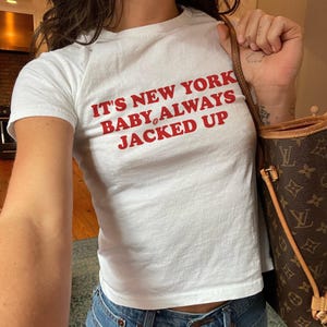 May include: A white short-sleeved t-shirt featuring red text that reads IT'S NEW YORK BABY, ALWAYS JACKED UP with a small coffee bean graphic between the words BABY and ALWAYS. The shirt has a crew neckline and a fitted silhouette.