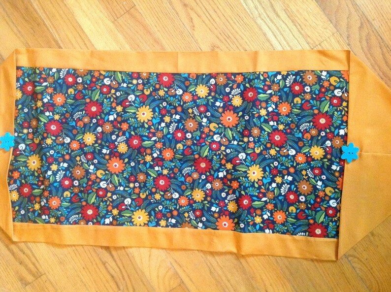 K&ouml;nnte beinhalten: Rechteckiger Tischl&auml;ufer mit floralem Muster in Rot, Orange, Gelb und Blau auf marineblauem Hintergrund. Mit einem orangefarbenen Rand und zwei blauen, bl&uuml;tenf&ouml;rmigen Verzierungen.