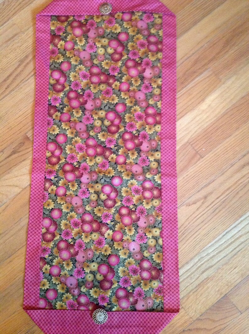 K&ouml;nnte beinhalten: Rechteckiger Tischl&auml;ufer mit floralem Muster, rosa Rand und einem Muster aus rosa und gelben Blumen. Der L&auml;ufer ist an jedem Ende mit dekorativen silbernen Kn&ouml;pfen verziert. Der Hintergrund ist ein Holzboden.