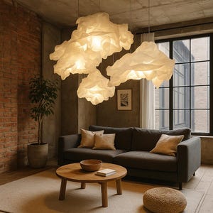 Può includere: Tre lampade a sospensione a forma di nuvola illuminano un soggiorno. Le luci sono bianche e pendono dal soffitto. Un divano grigio scuro con cuscini beige si trova sotto una grande finestra. Nella stanza ci sono anche un tavolino rotondo in legno e una pianta in vaso.