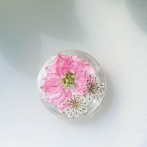 Puede incluir: Un objeto redondo y transparente de resina que contiene una flor rosa y pequeñas flores blancas. El arreglo floral está centrado dentro de la resina transparente, creando una pieza decorativa. El fondo es una superficie blanca con sombras.